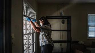 Gil Barel moves the blinds up to let in more light into her living room in Pasadena on Aug. 2, 2026. Barel is currently not living in her apartment because of the Eaton Fire of January 2025.