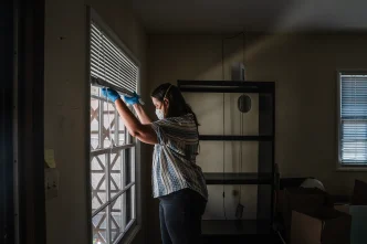 Gil Barel moves the blinds up to let in more light into her living room in Pasadena on Aug. 2, 2026. Barel is currently not living in her apartment because of the Eaton Fire of January 2025.